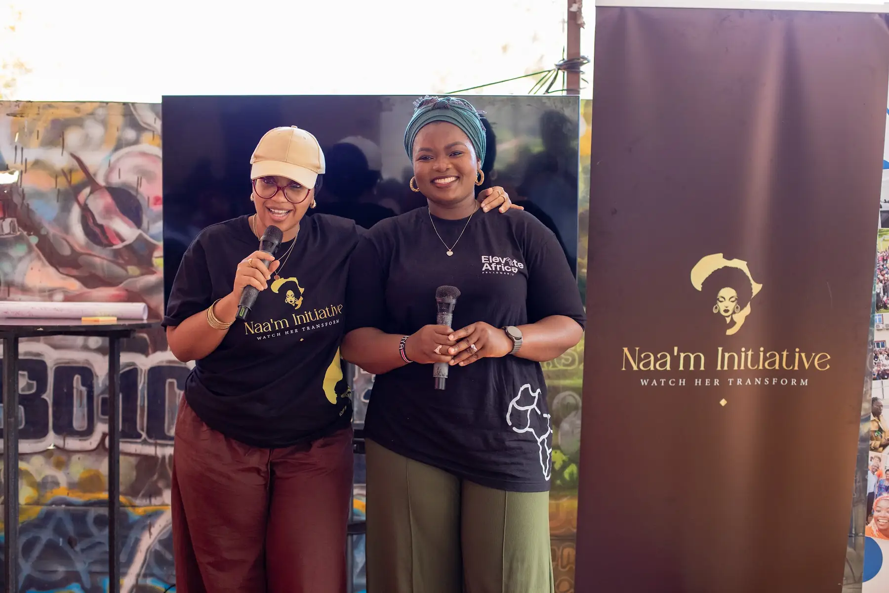 Two speakers sharing a microphone at the NAA’M Initiative banner